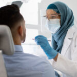 Islamic Dentist Lady In Medical Mask And Face Shield Checking Male Patient's Teeth With Sterile Tool, Muslim Female Stomatologist Doctor In Hijab Making Dental Treatment To Young Man At Modern Clinic