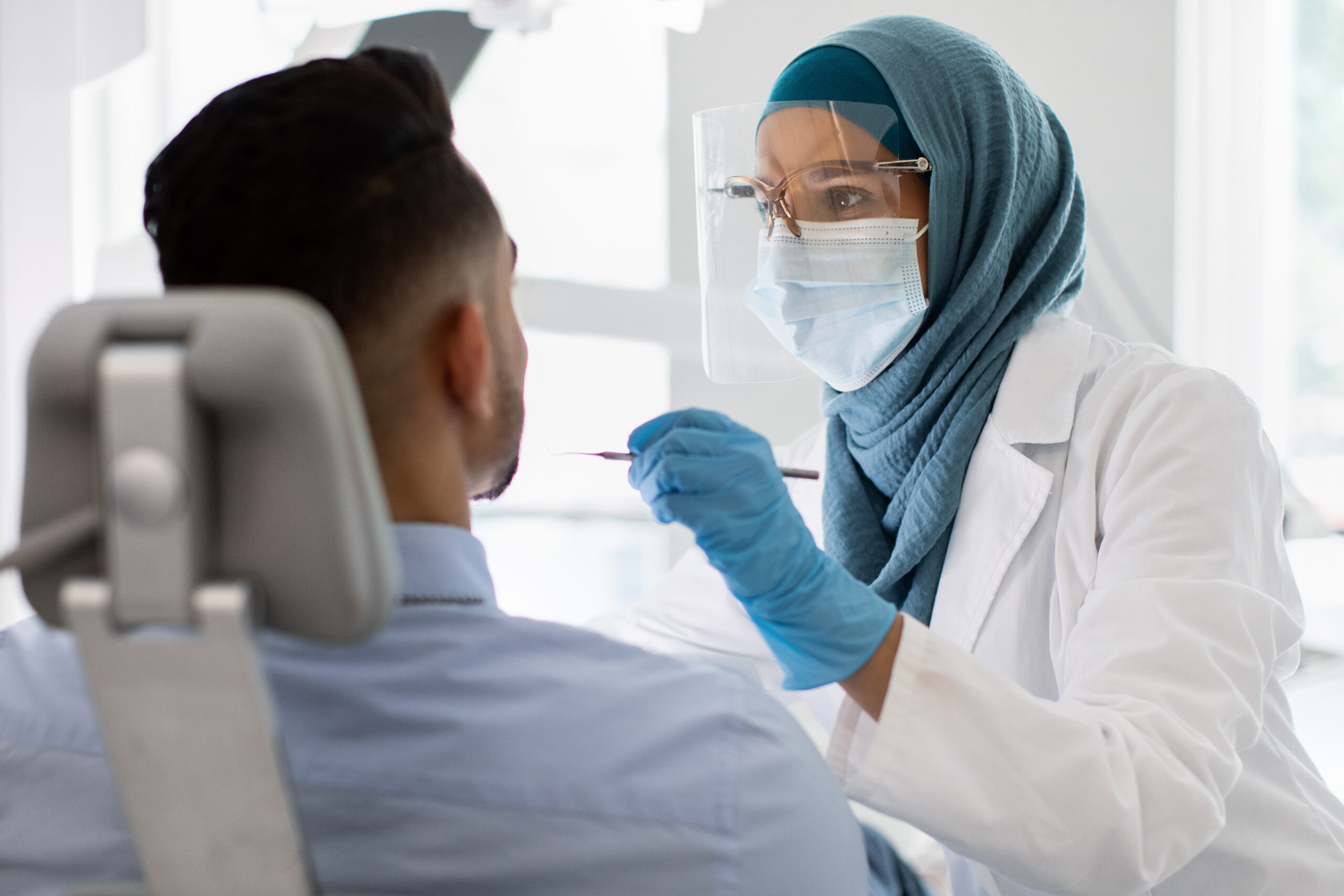 Islamic Dentist Lady In Medical Mask And Face Shield Checking Male Patient's Teeth With Sterile Tool, Muslim Female Stomatologist Doctor In Hijab Making Dental Treatment To Young Man At Modern Clinic