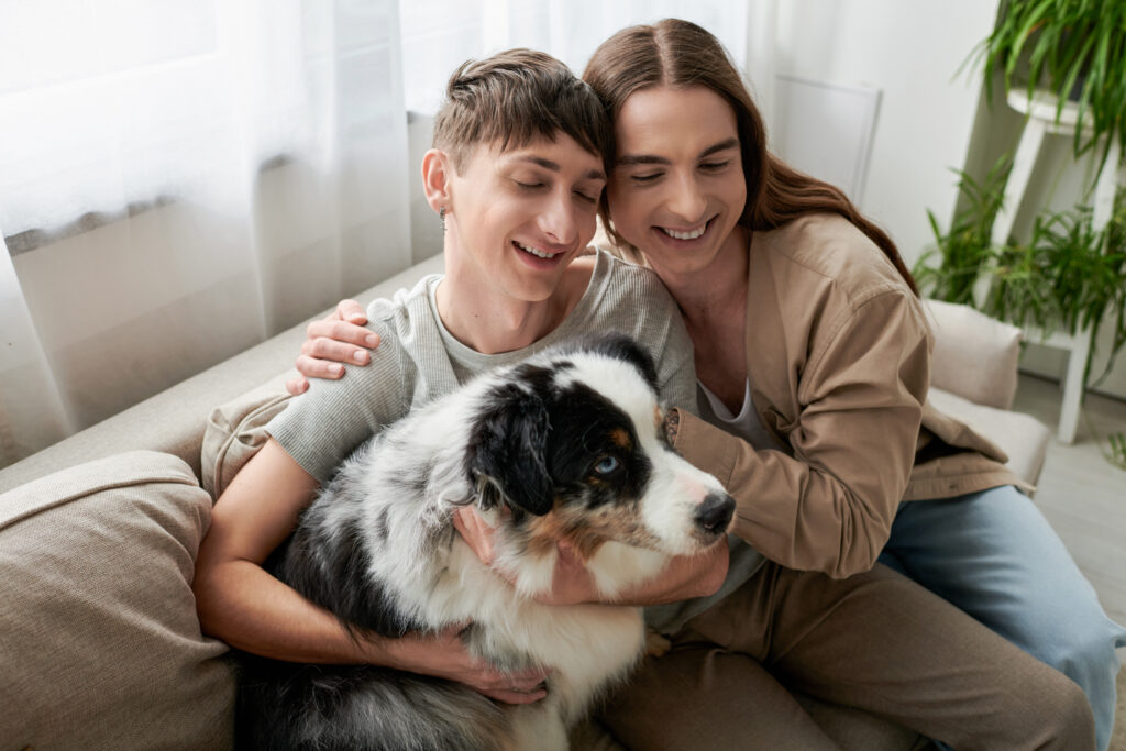 High angle view of smiling homosexual couple with closed eyes hugging near Australian shepherd dog on couch in modern living room at home