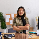 successful happy asian clothes designer with folded arms looking at camera near work desk in studio