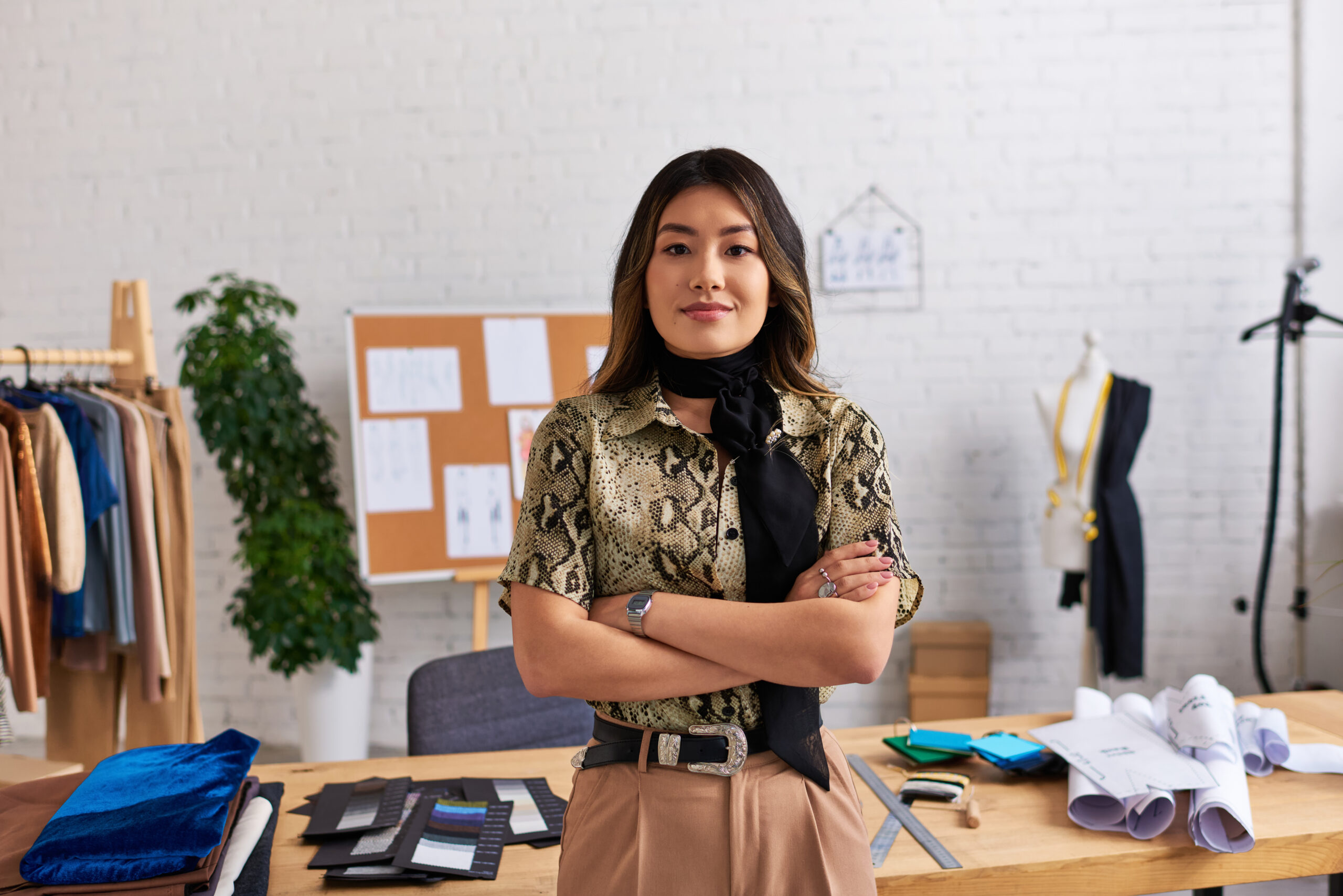 successful happy asian clothes designer with folded arms looking at camera near work desk in studio